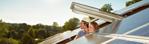 PIA Zwei Personen, schauen aus einem Dachfenster heraus auf ein Dach mit Solarpaneelen. Im Hintergrund sind Bäume und ein klarer Himmel zu sehen.